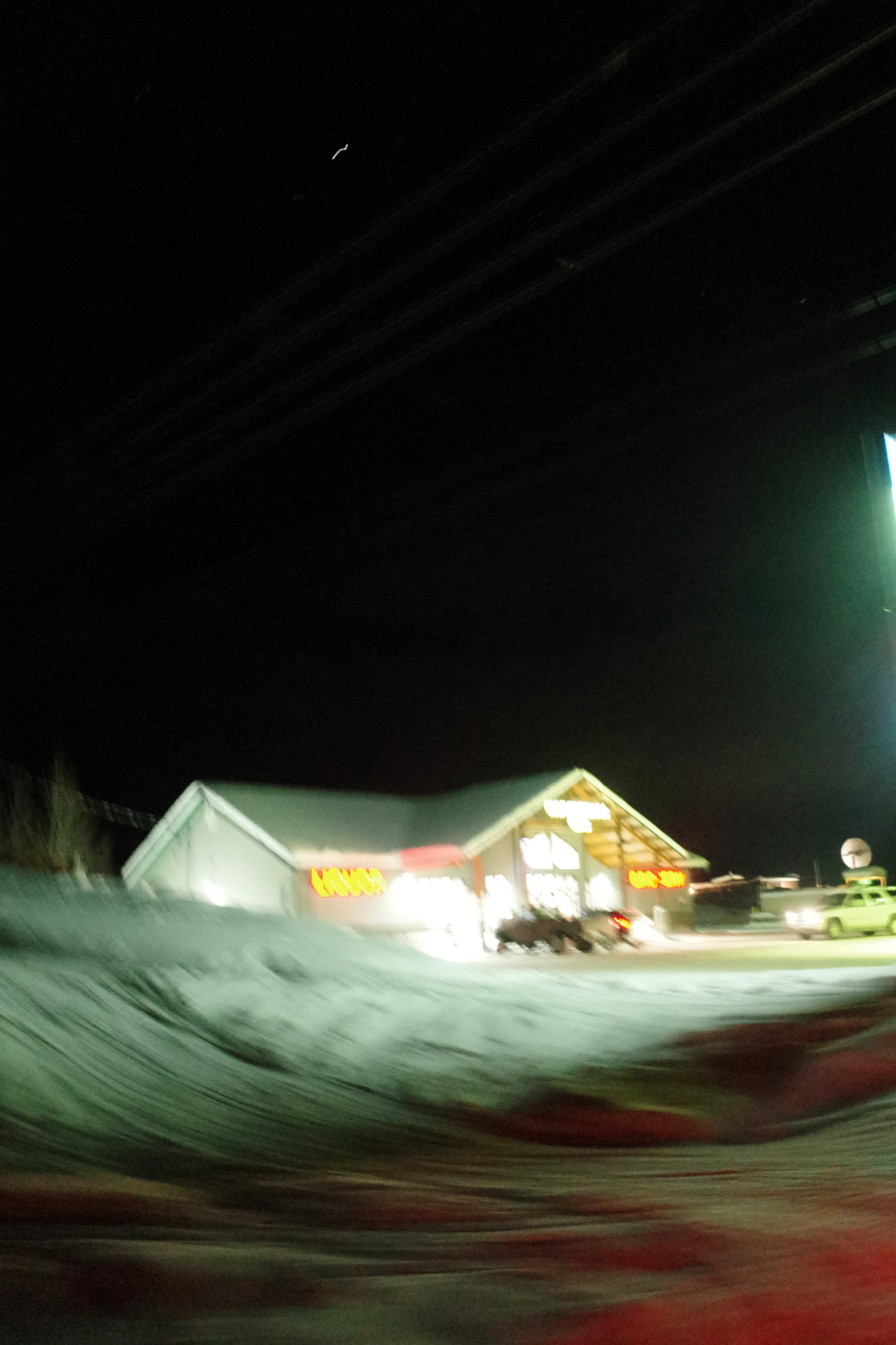 A snowbound roadside building and parked cars blur beneath bright neon lights at night.