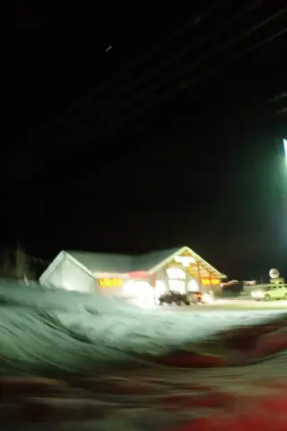 A snowbound roadside building and parked cars blur beneath bright neon lights at night.
