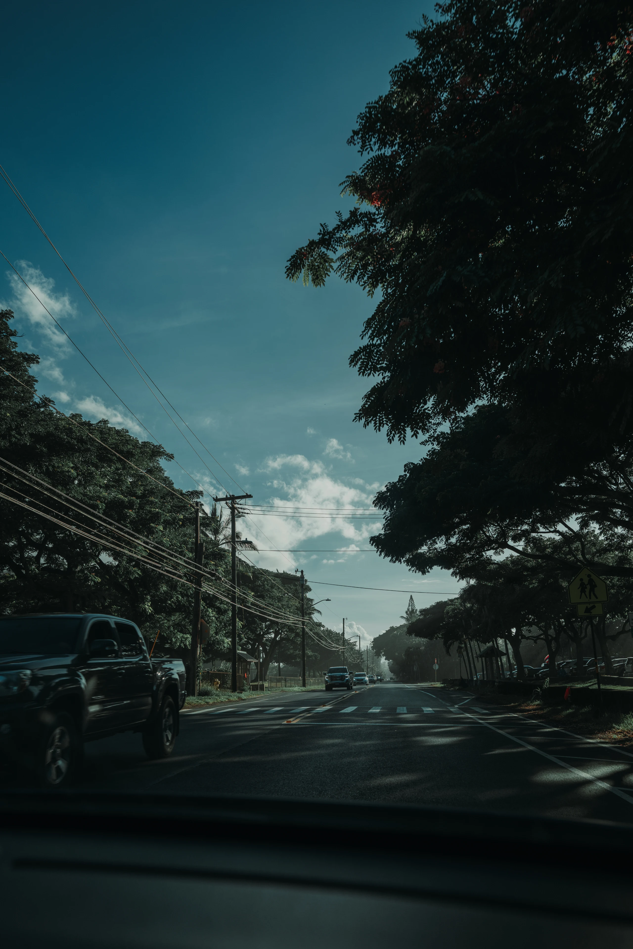 A sunlit O‘ahu road seen through a windshield, with a pickup passing beneath large trees, utility wires, and a deep blue sky.