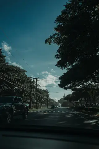 A sunlit O‘ahu road seen through a windshield, with a pickup passing beneath large trees, utility wires, and a deep blue sky.