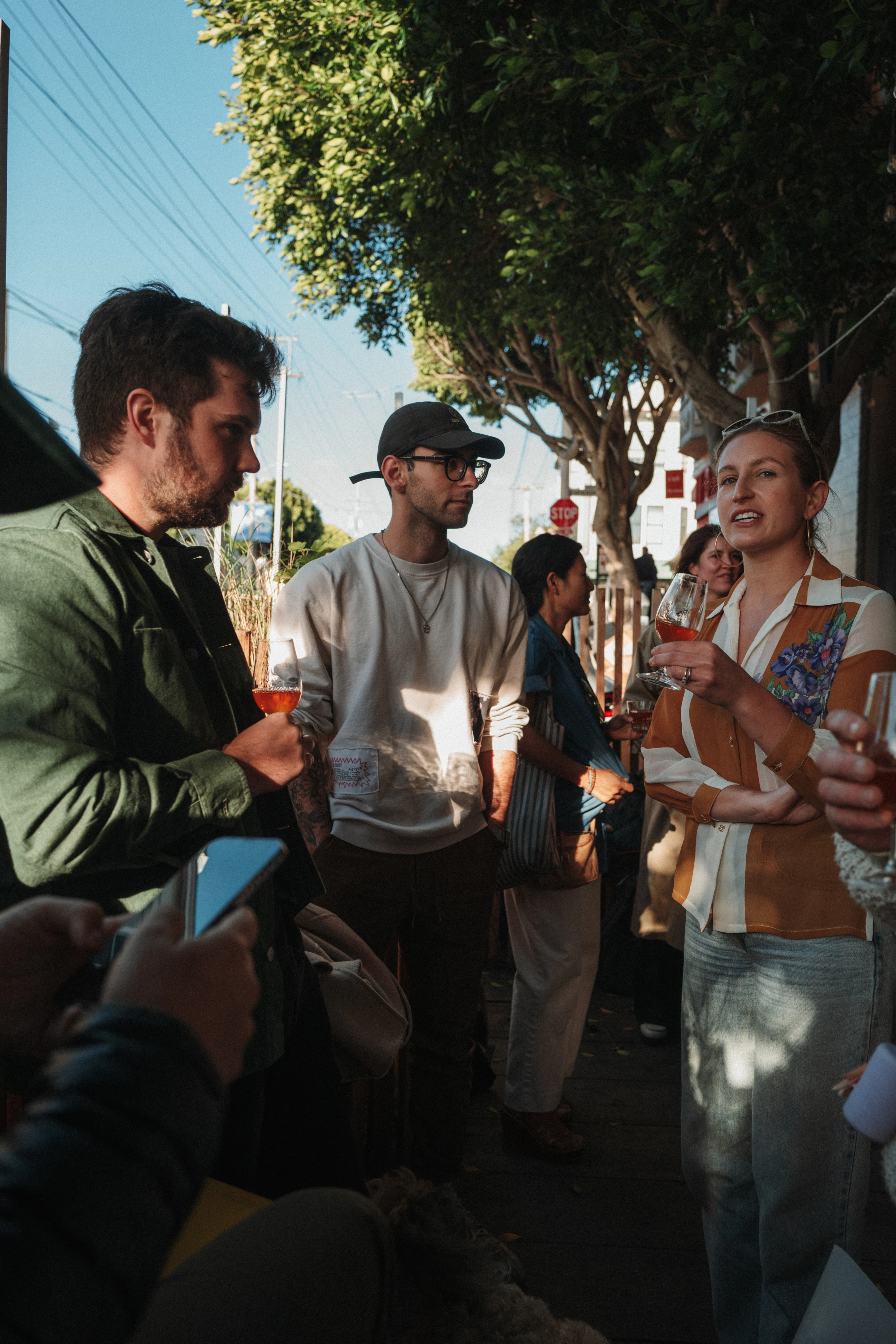 Three people hold wine glasses and talk on a shaded sidewalk beneath trees, with other guests behind them.