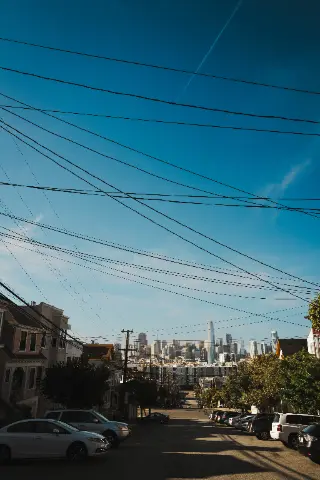 A steep San Francisco residential street beneath many utility wires with downtown towers in the distance.
