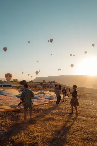 Hot-air balloons float over Cappadocia at sunrise while people prepare on the launch field below.