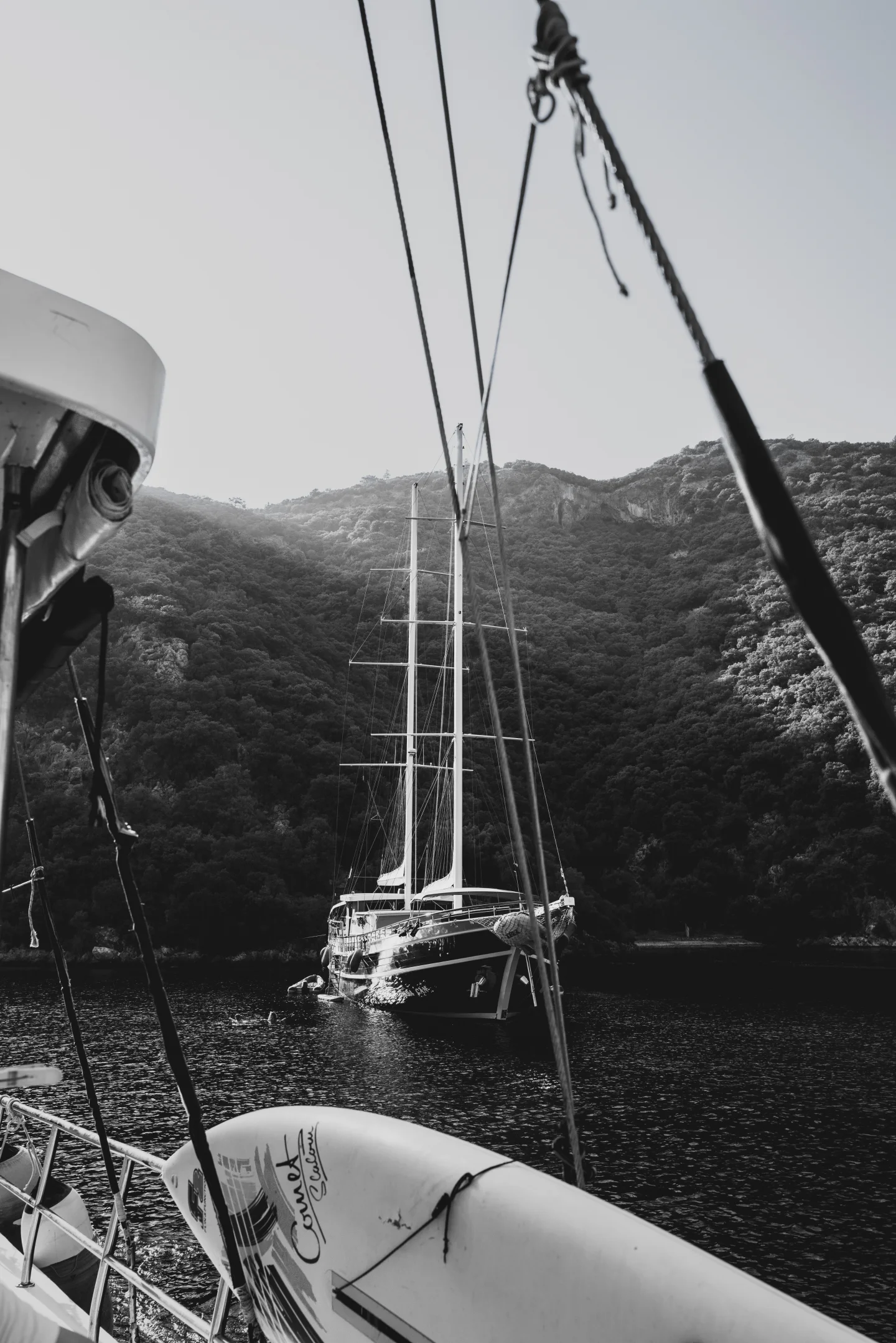 A monochrome view across a tender toward an anchored sailboat and steep hills.