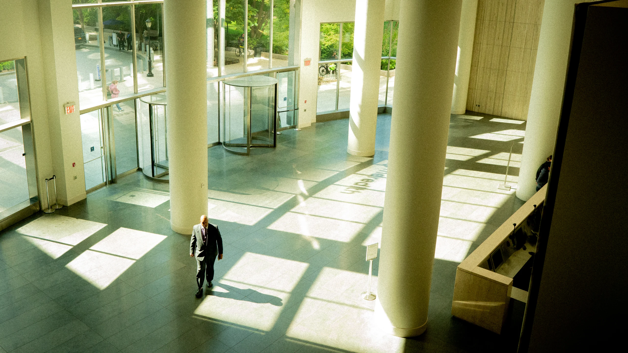 A suited man walks across a tall lobby where broad columns and windows cast a geometric grid of sunlight and shadow.