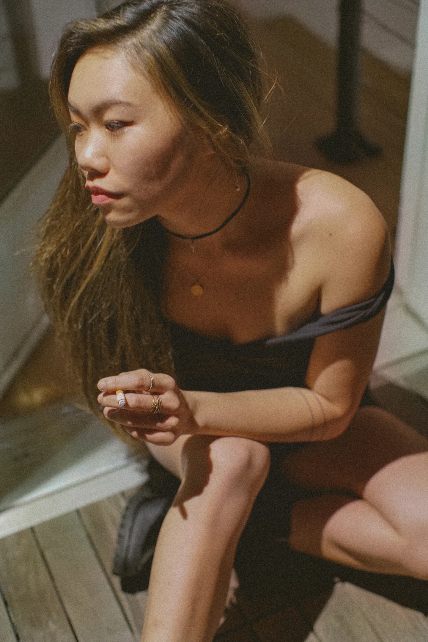A woman in a dark off-shoulder dress sits beside a window, looking left while holding a cigarette in angular sunlight.