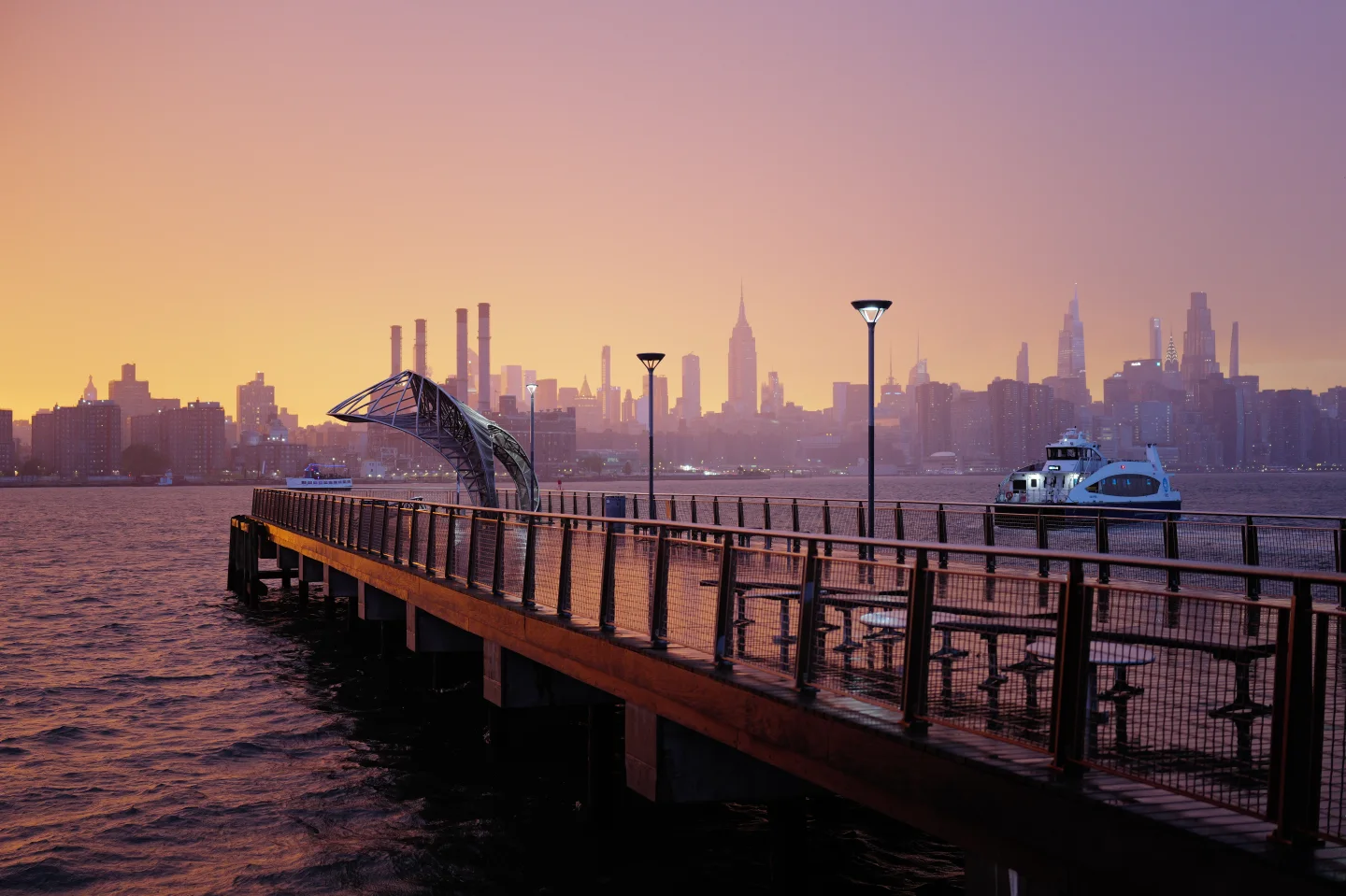 Williamsburg waterfront pier and ferry with the Manhattan skyline beneath a pink and violet dusk sky.