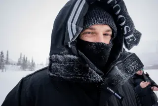 A bundled person looking toward the camera with snow collecting on a black hood and face covering.