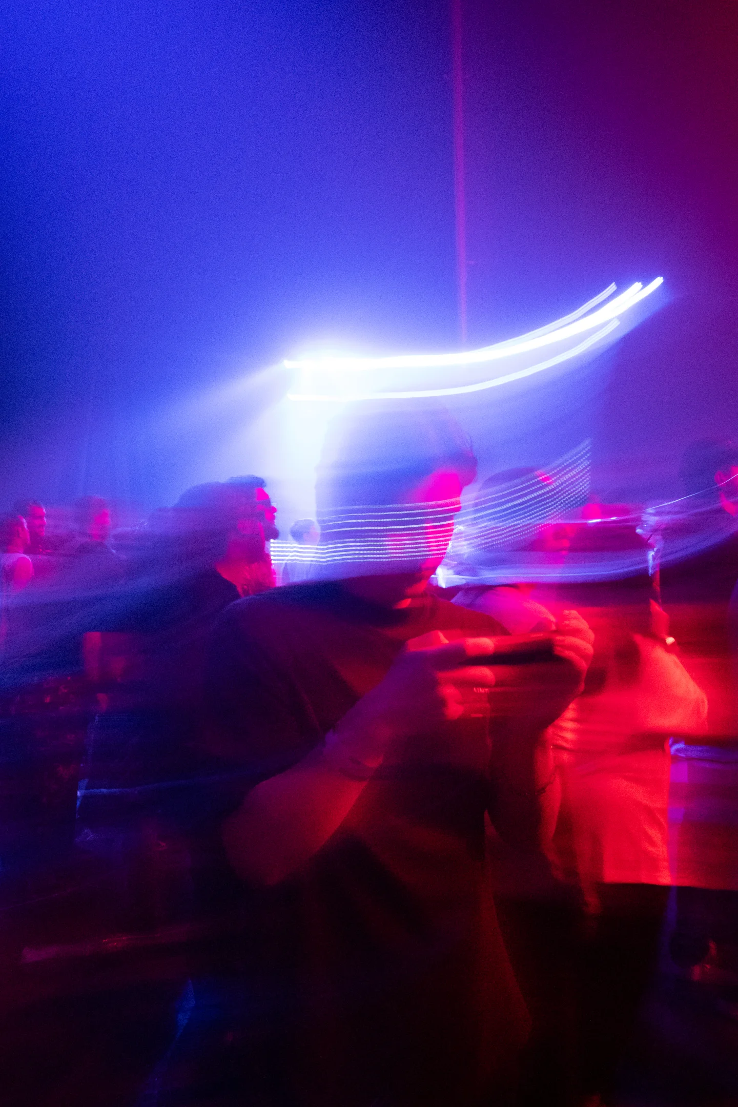 A blurred white light streak above a concert crowd in red and violet light.