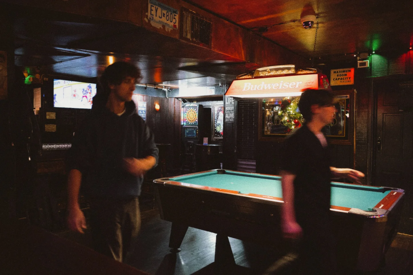 Friends gather among several pool tables in a dark neighborhood hall.