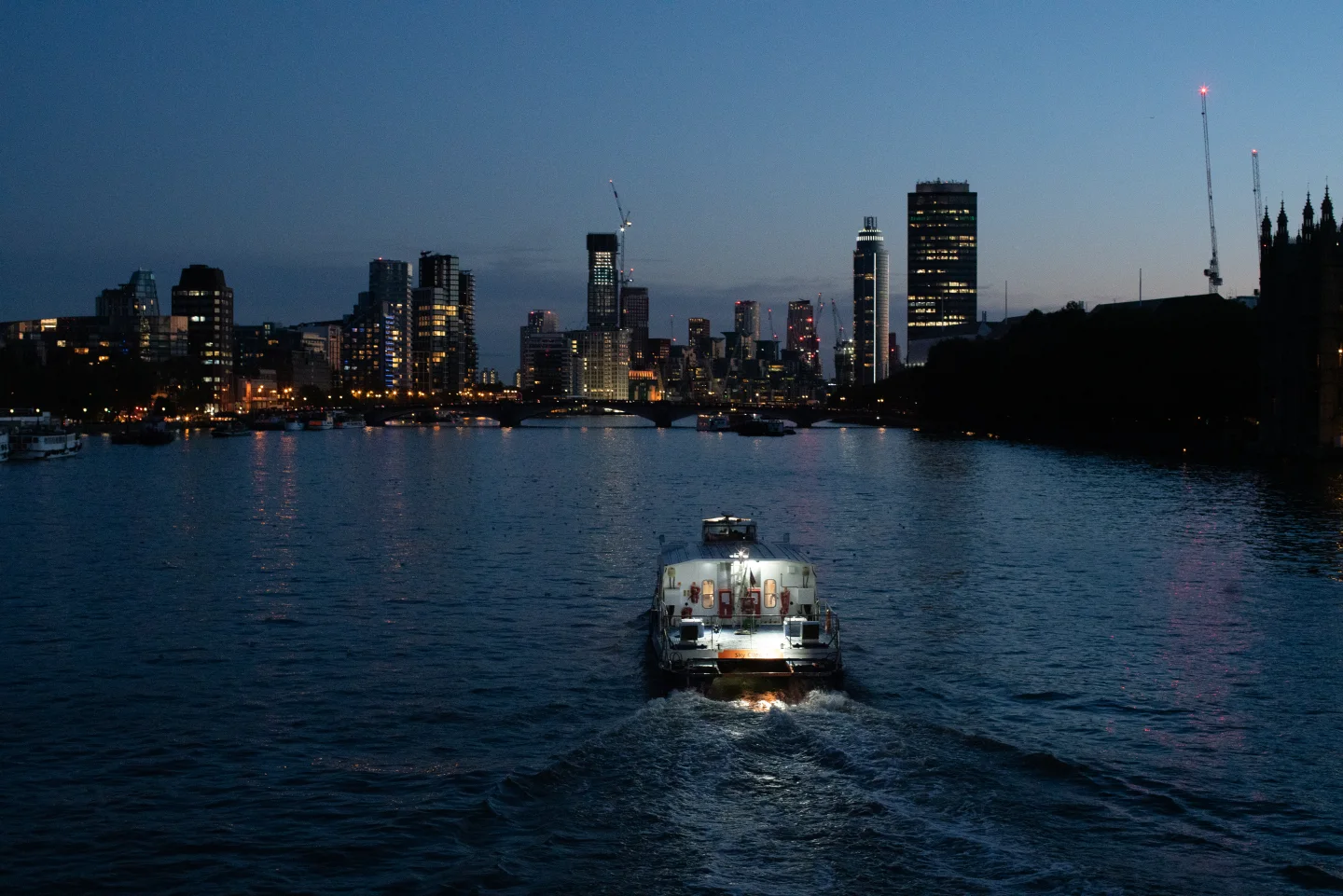 A passenger boat approaches on the Thames with the evening skyline beyond.