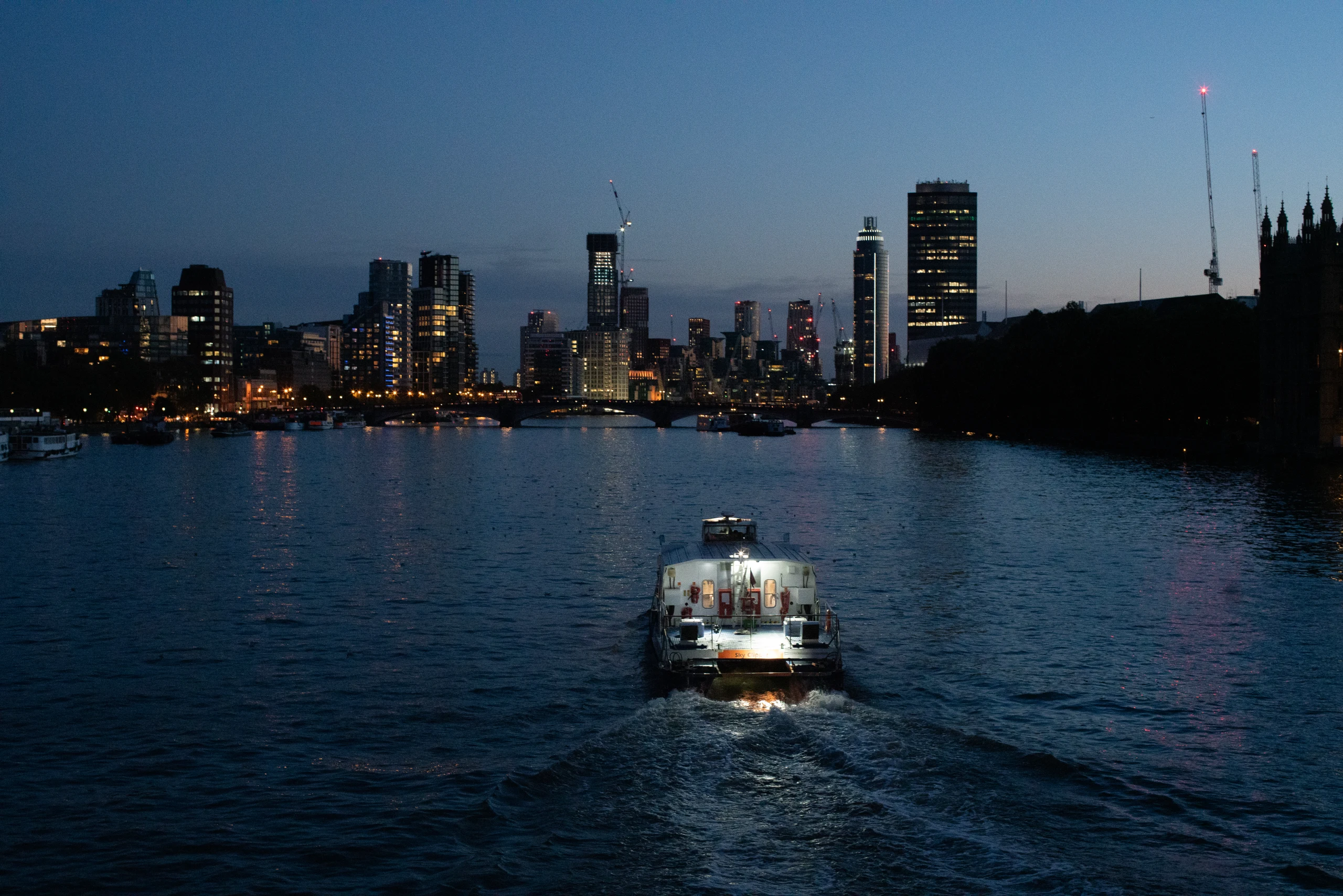 A passenger boat approaches on the Thames with the evening skyline beyond.