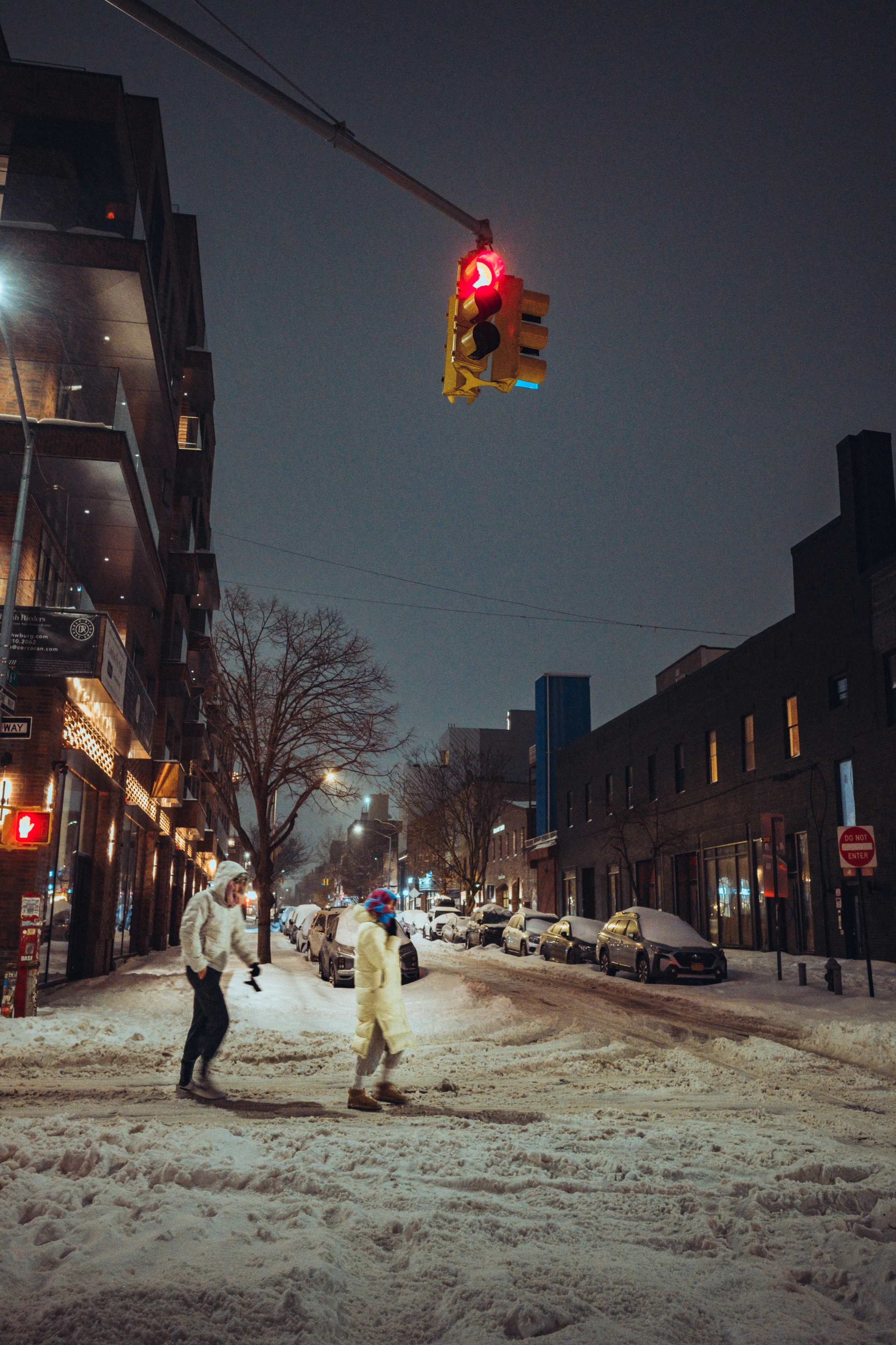 Two people crossing a snow-covered Brooklyn street under a red light