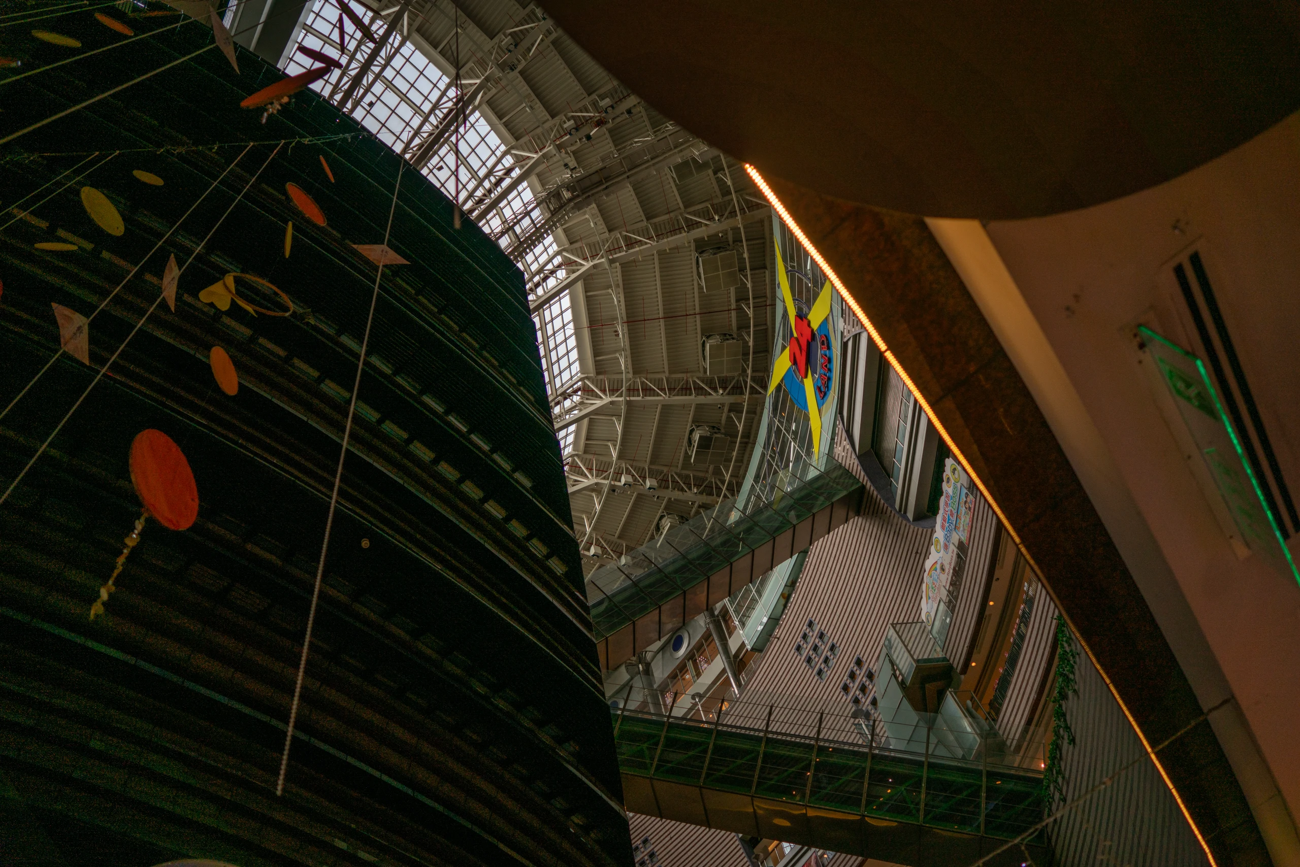 Upward tilted view inside a Taipei atrium with curved dark walls, suspended shapes, walkways, and a glass-and-steel roof.