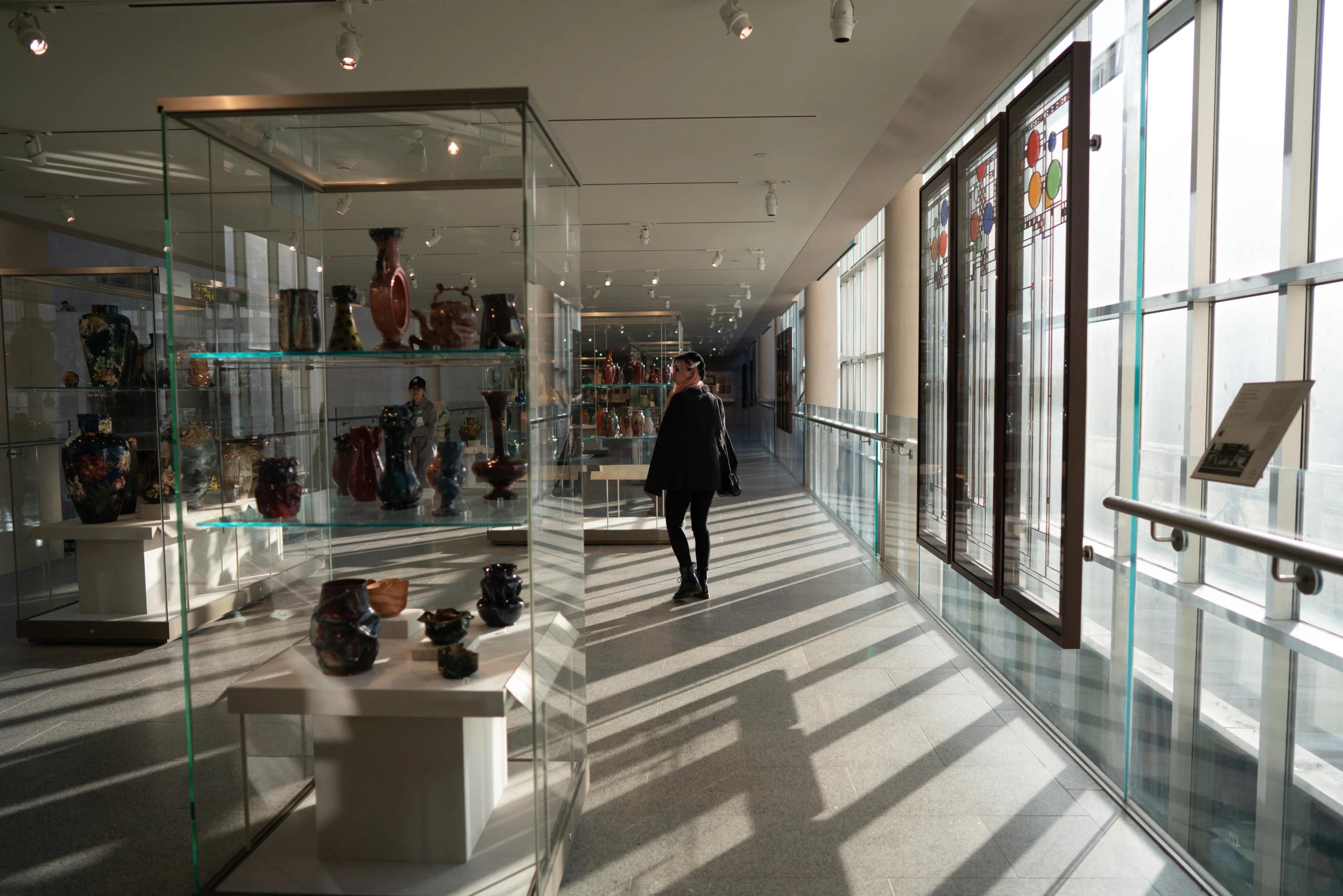 A bright museum gallery corridor with sculptures, windows, and one walking figure.