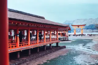 A red shrine corridor stands beside an exposed tidal flat, with a large torii gate and visitors beyond.