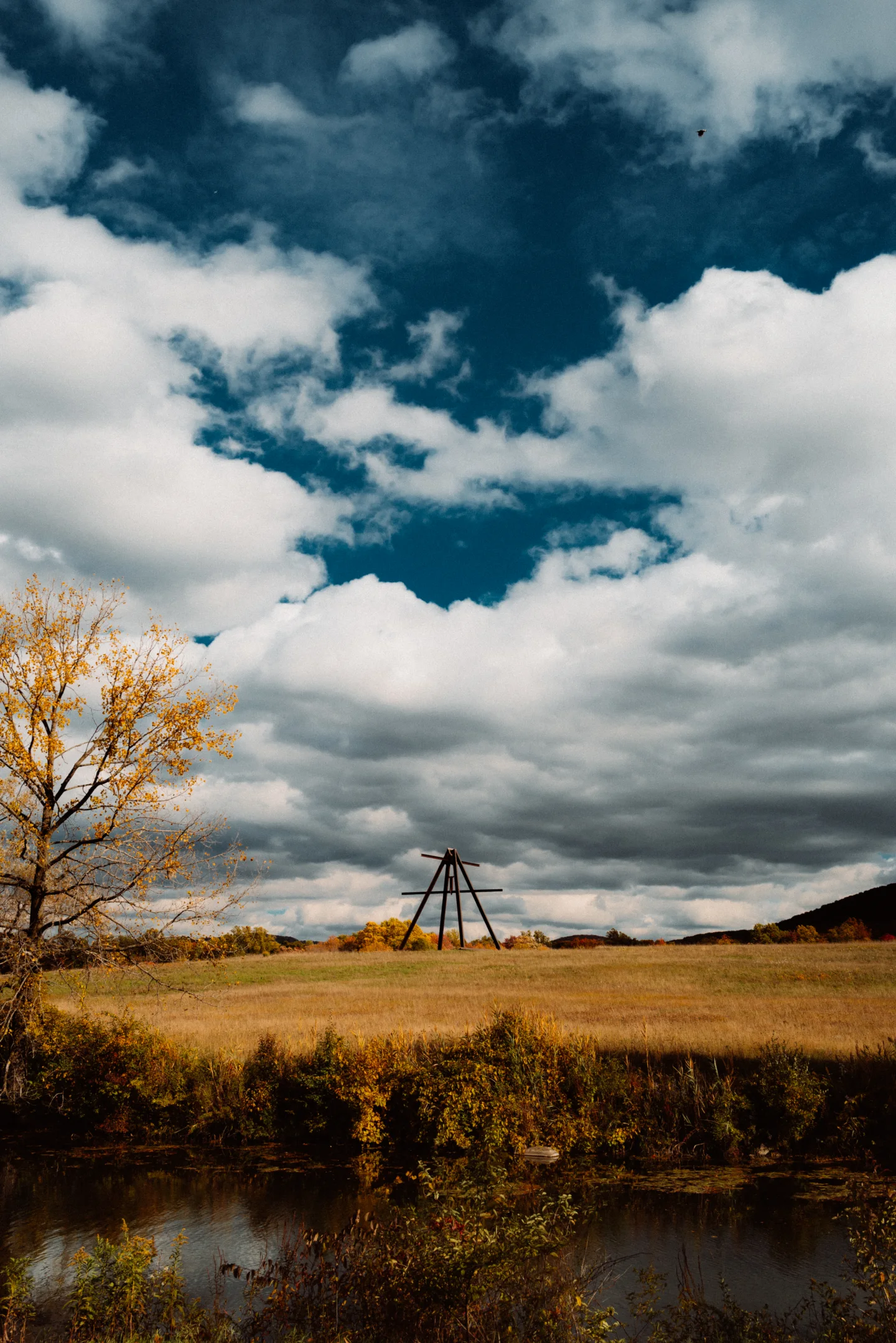 A dark steel sculpture stands in an autumn field beneath towering blue-and-white clouds, with a yellow-leaved tree beside a narrow pond.