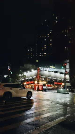 Junior’s Restaurant glows red and white on a rainy New York night as a white vehicle crosses the wet intersection.