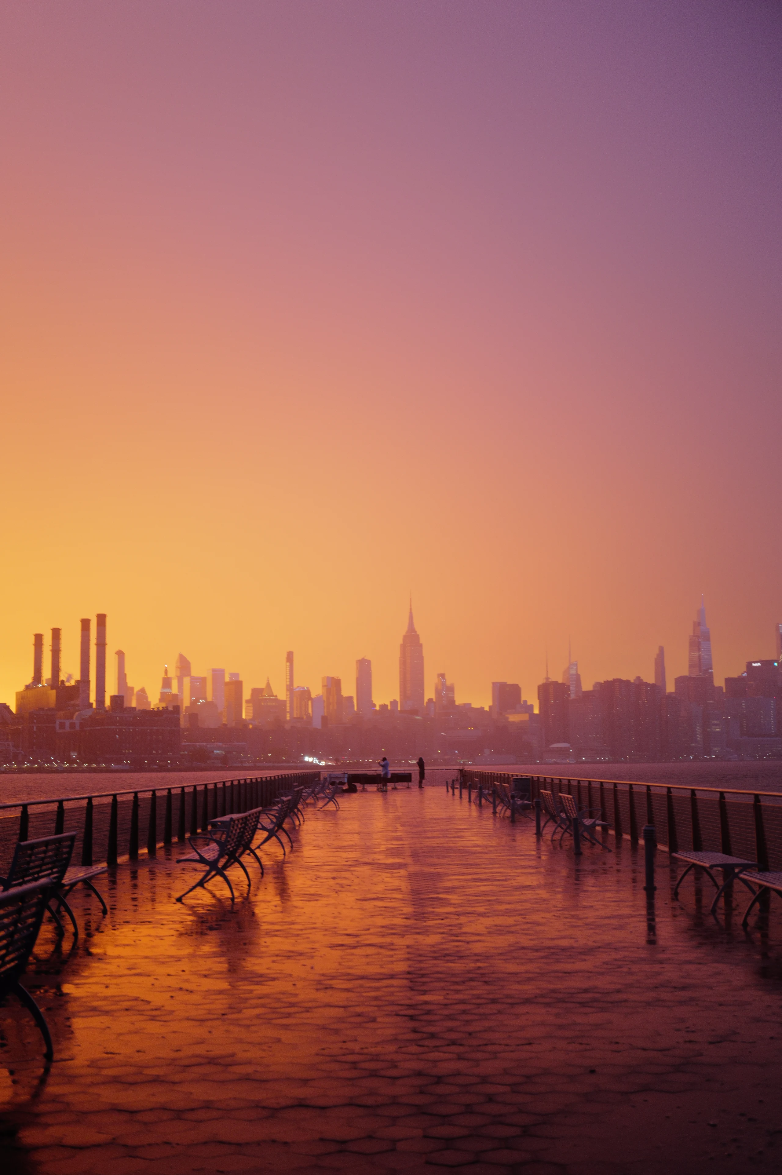 Empty waterfront pier with benches reflecting a golden sunset and the Manhattan skyline.