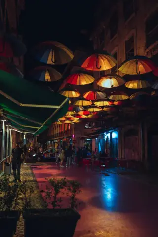 Colorful umbrellas hang above a restaurant-lined lane with people, outdoor tables, and a blue reflection on the pavement.