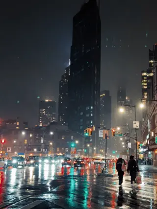 Cars and pedestrians move through a rain-soaked Manhattan avenue at night, with red and green traffic lights reflected across the pavement.