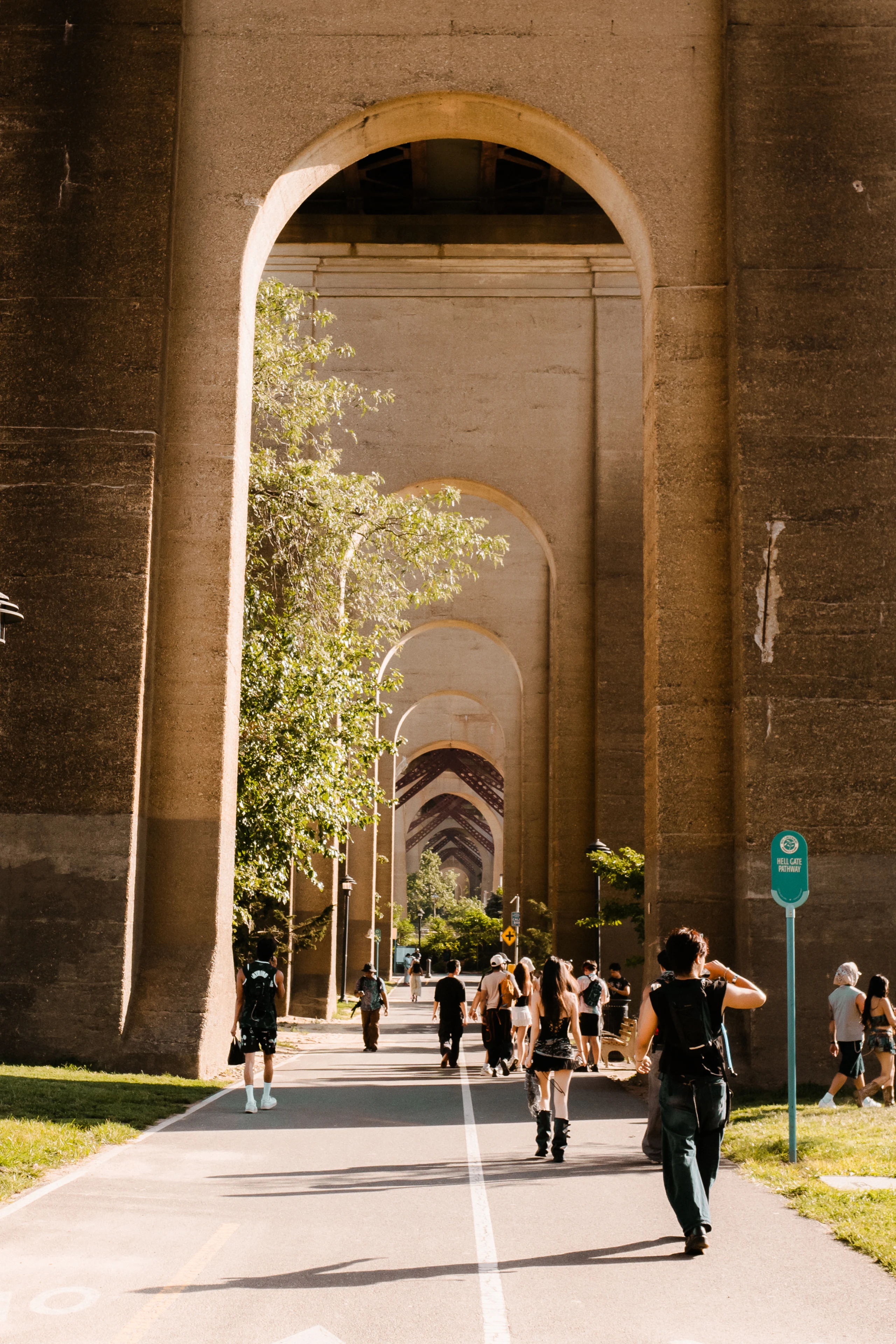 Pedestrians walk along a sunlit path beneath a repeating line of tall concrete bridge arches.