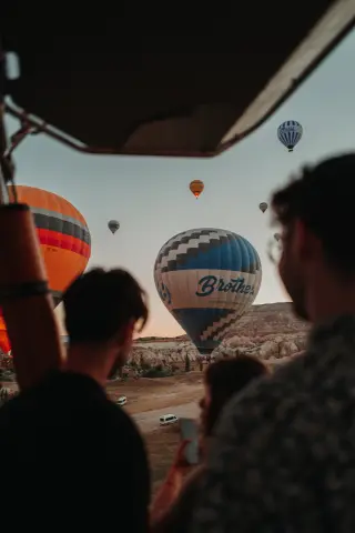 Silhouetted passengers look from a hot-air balloon basket toward a large blue-and-white balloon descending over Cappadocia.