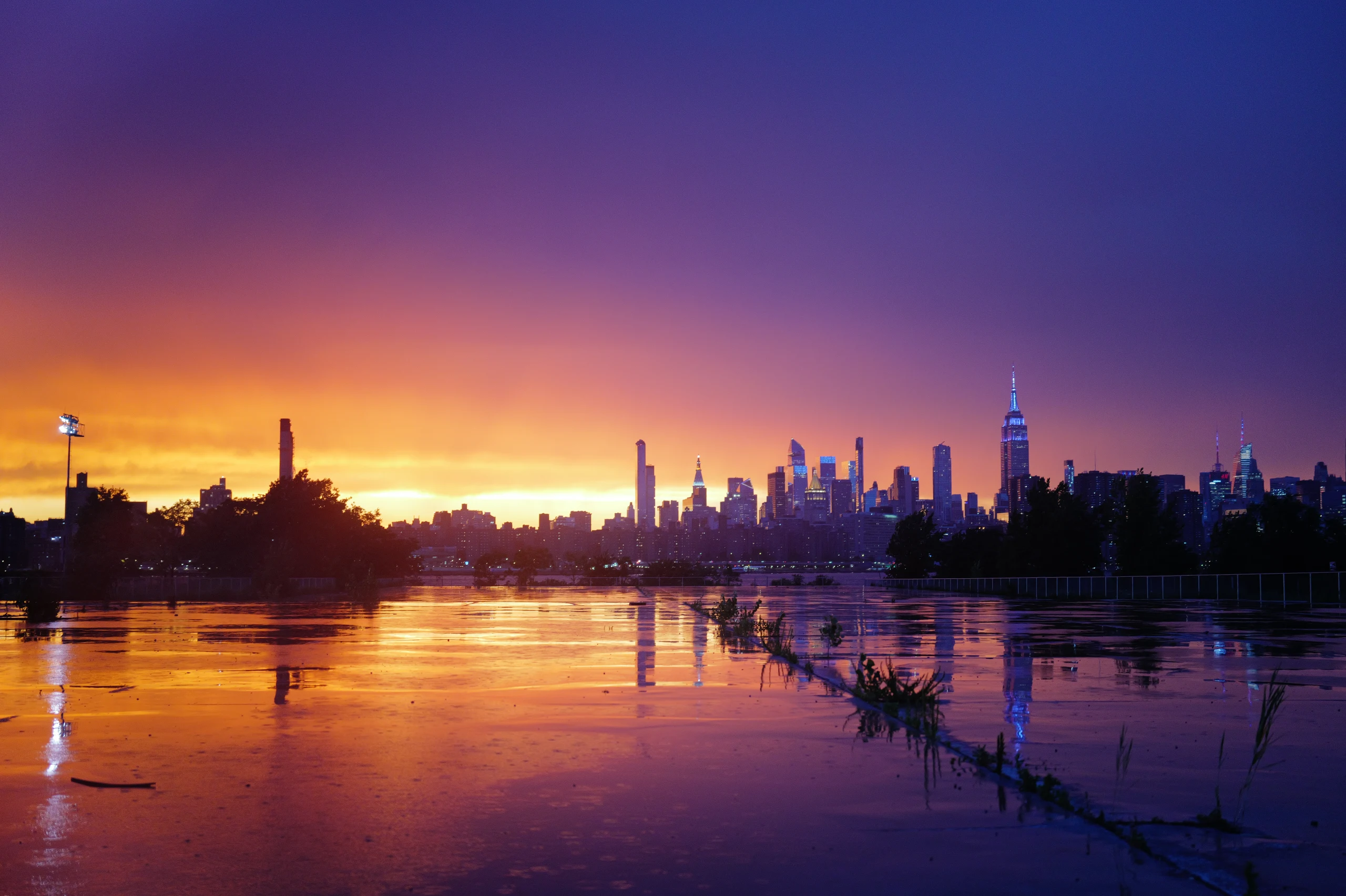 Manhattan skyline beneath an orange and violet storm sky reflected across a wet field.