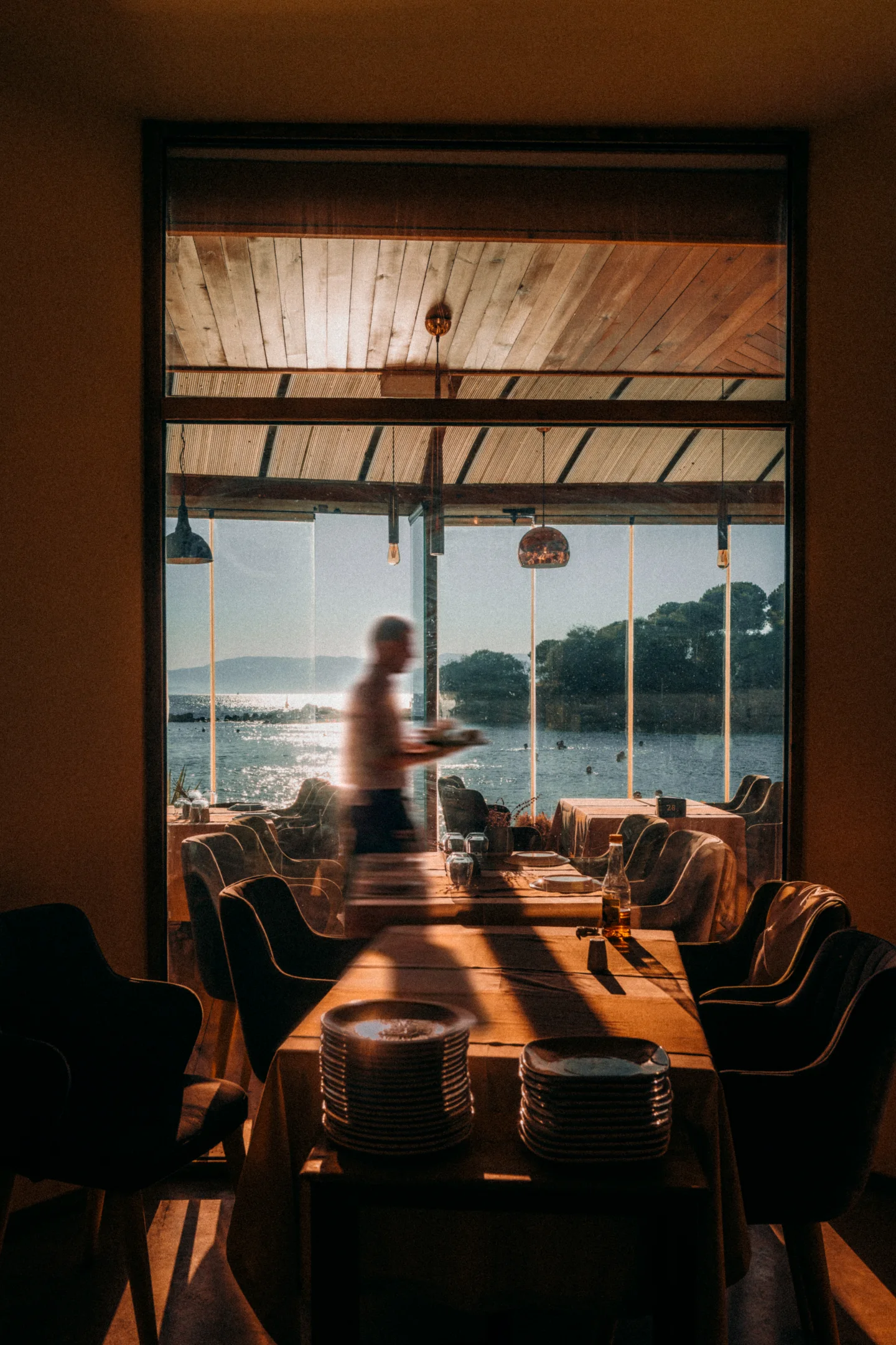 A blurred figure crosses a sunlit waterfront dining room, seen through a dark interior doorway.
