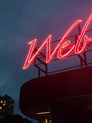 A large red neon sign reading Web glows above a dark curved building against a blue-gray evening sky.