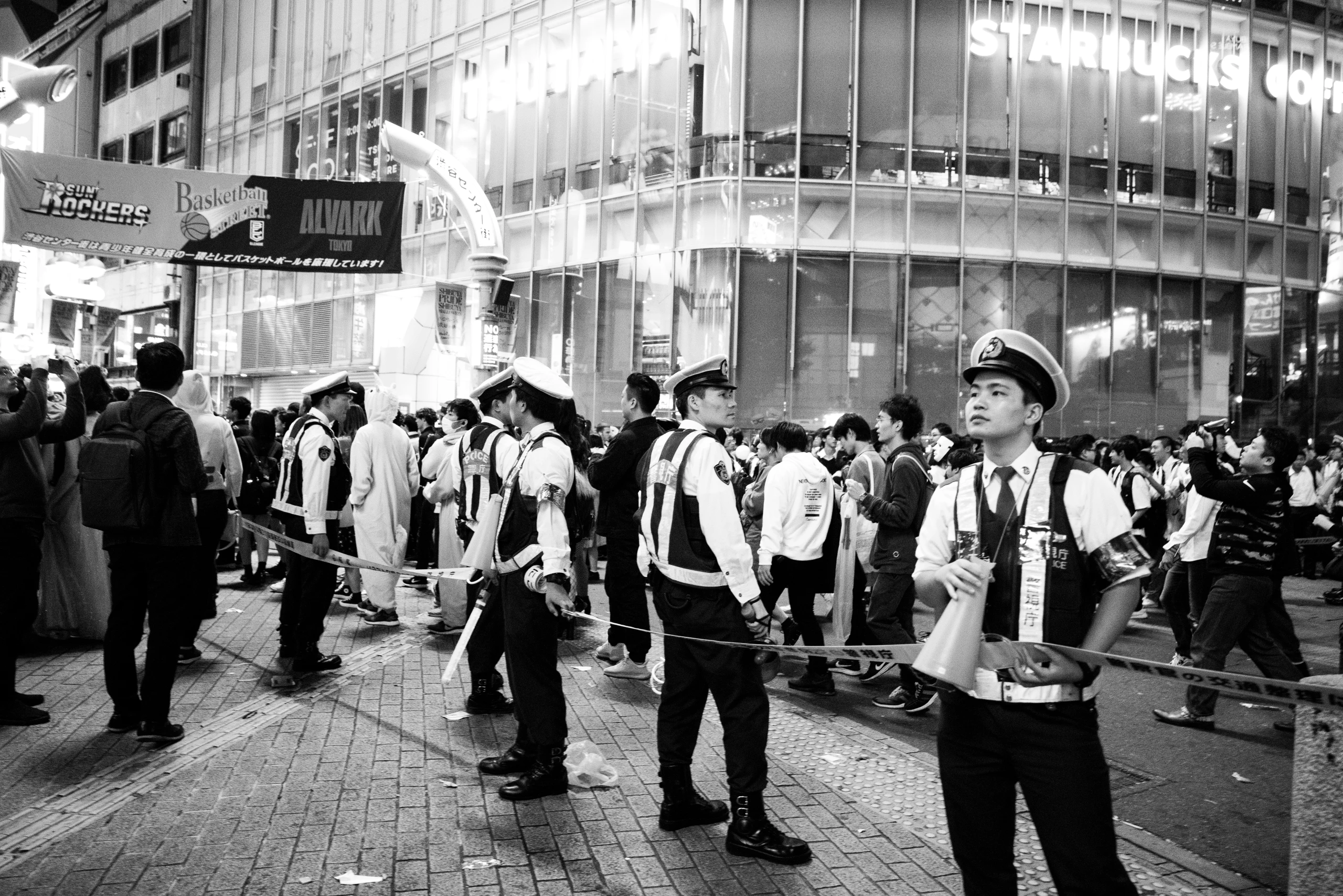 Uniformed police hold a cordon through a crowded Tokyo street at night beneath bright storefront lights.