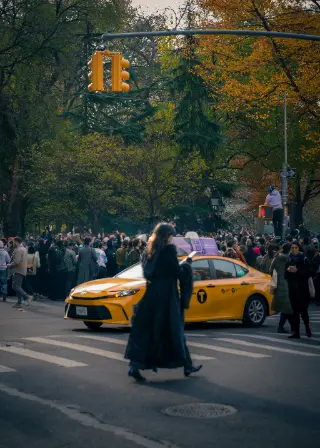 A person in a long dark coat crosses beside a yellow taxi in autumn.