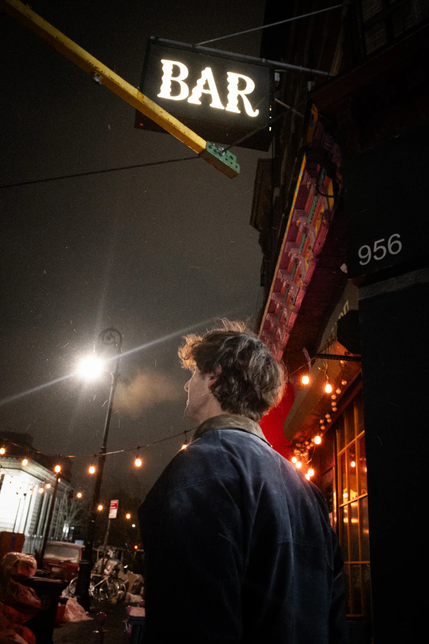 A man looks up beneath a bright BAR sign on a Williamsburg sidewalk.