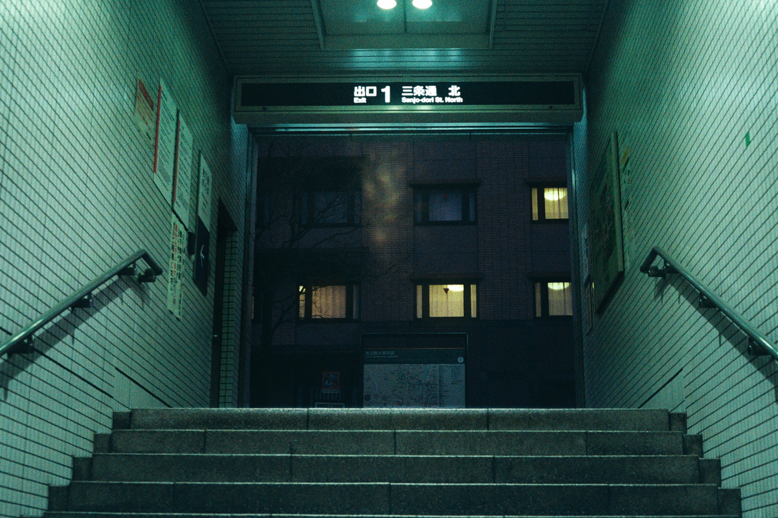 A short stairway rises between green tiled walls toward an exit sign and a dark building beyond.
