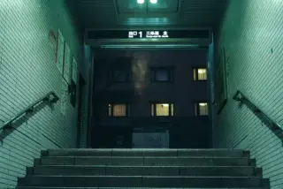 A short stairway rises between green tiled walls toward an exit sign and a dark building beyond.