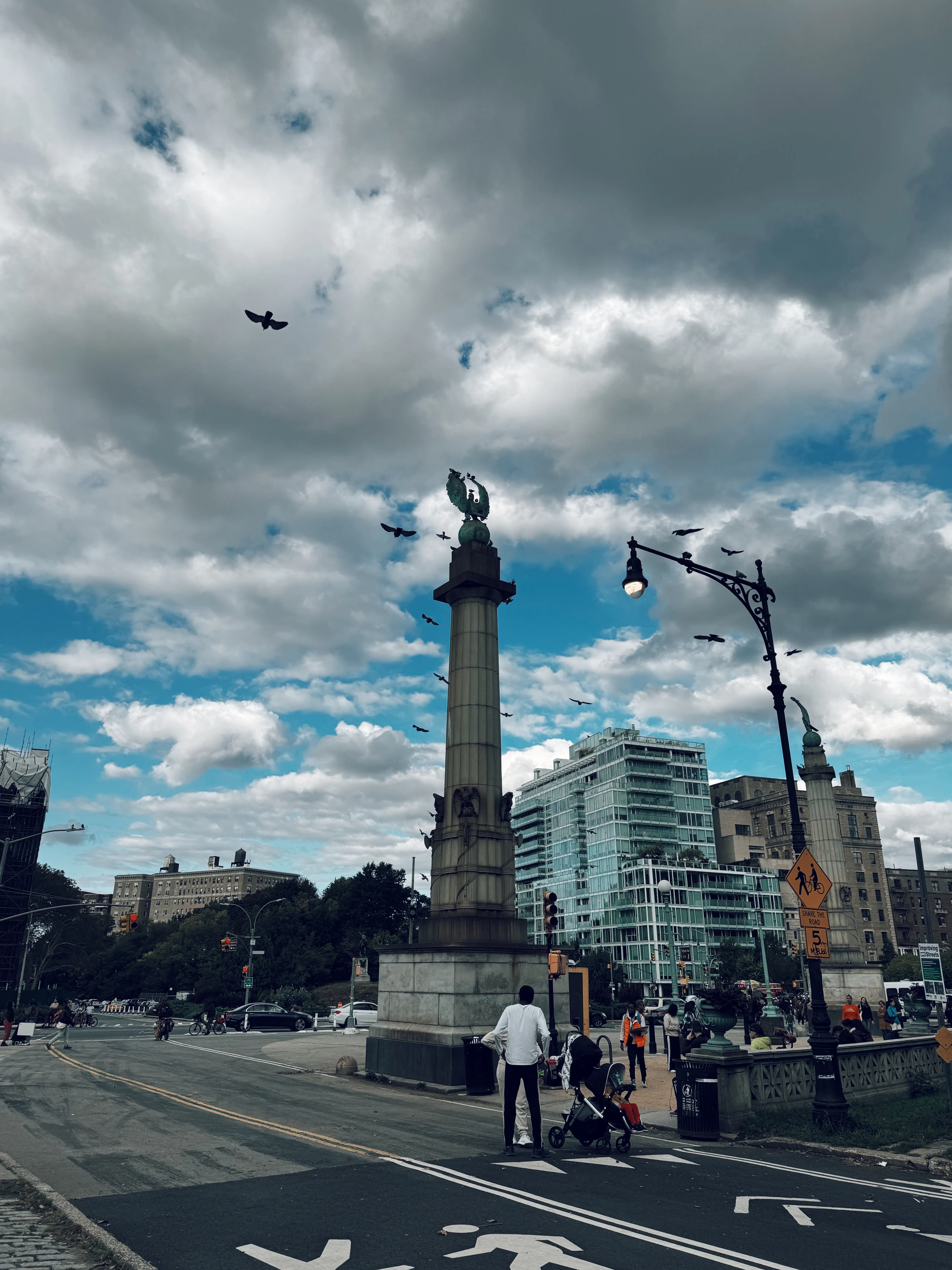 Pigeons fly around a tall stone monument and ornate streetlamp beneath layered gray-and-blue clouds at a Manhattan plaza.