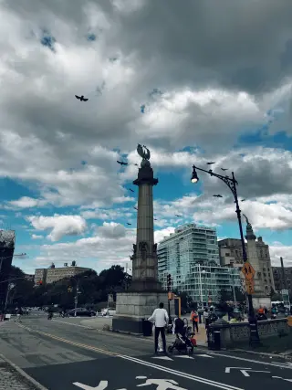 Pigeons fly around a tall stone monument and ornate streetlamp beneath layered gray-and-blue clouds at a Manhattan plaza.