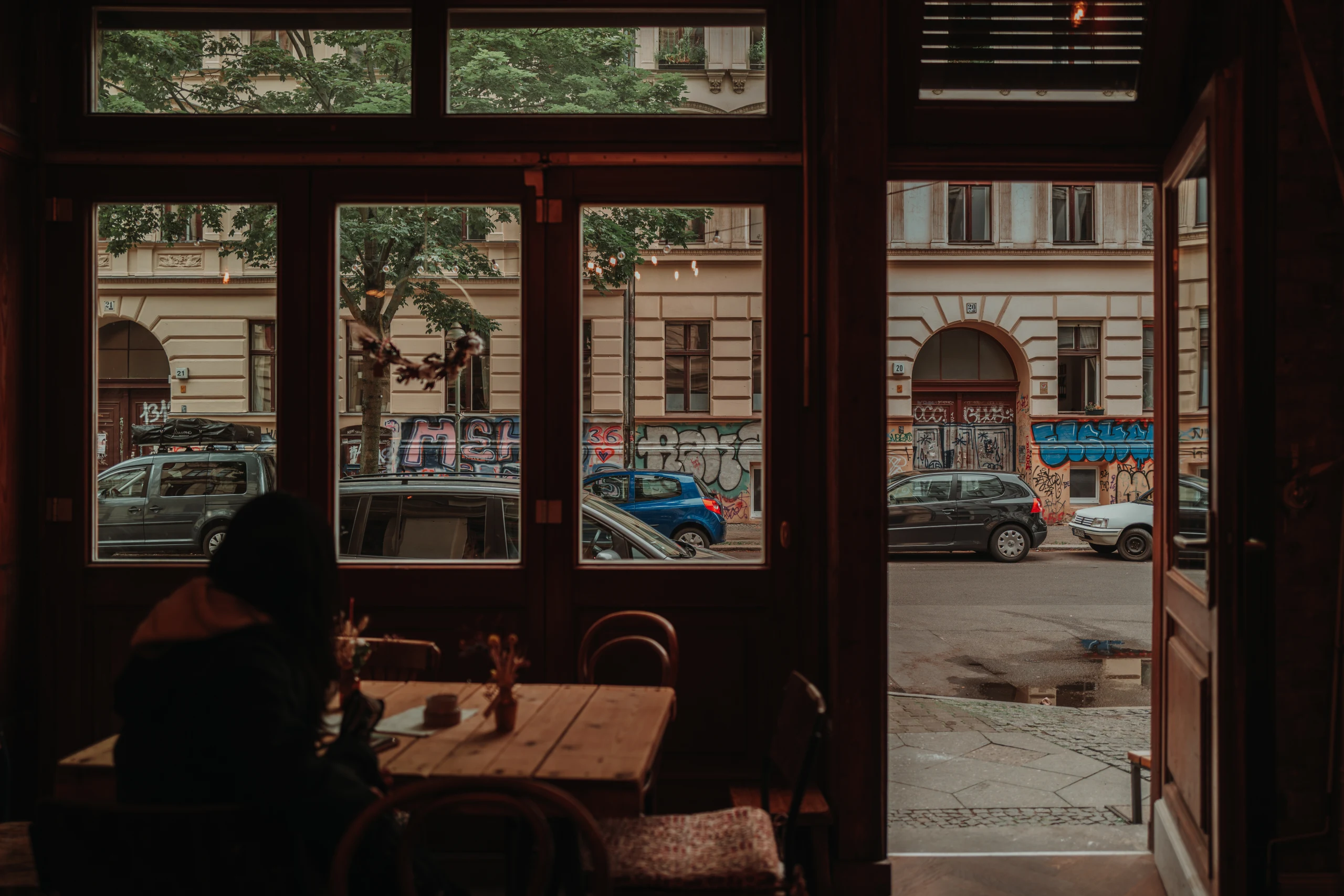 View through a dark café interior toward parked cars and graffiti-covered buildings in Berlin.
