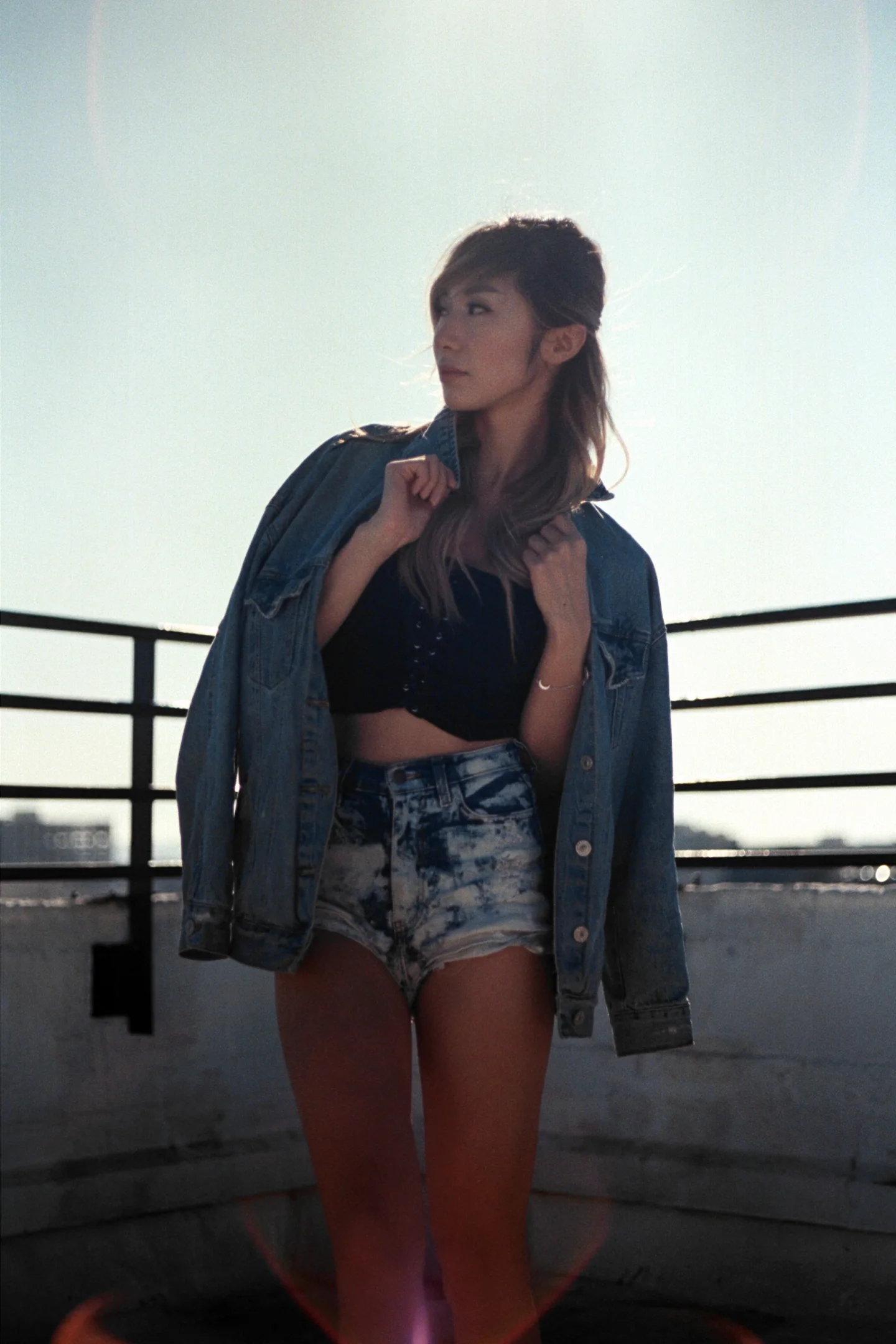 A woman in a jacket and shorts standing at a rooftop railing in bright backlight.
