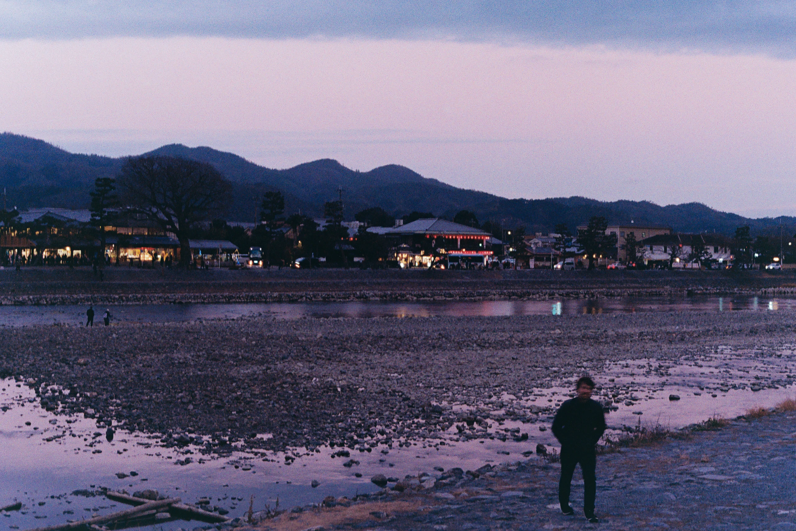 A lone person stands on a broad tidal shore at dusk.