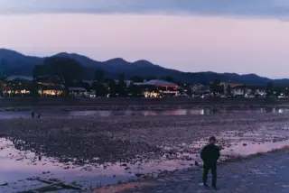 A lone person stands on a broad tidal shore at dusk.