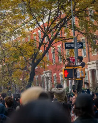 A crowd walking beneath autumn trees at a New York street corner.