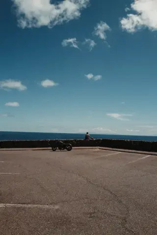 A motorcycle and a lone figure stand at an asphalt overlook facing the blue sea and sky.