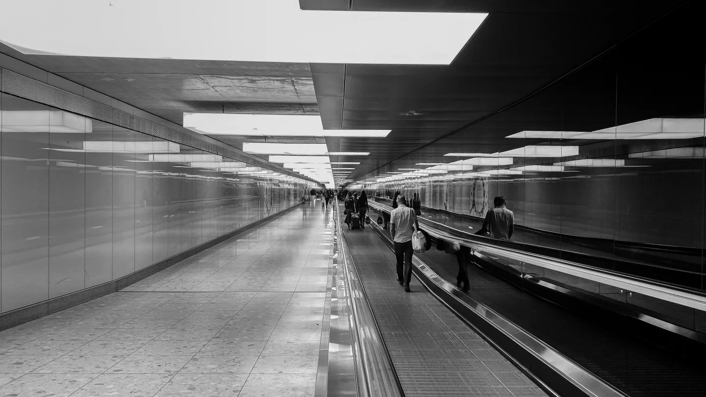 Black-and-white photograph of travelers on an airport walkway, bordered by glass and mirrored panels beneath rectangular ceiling lights.