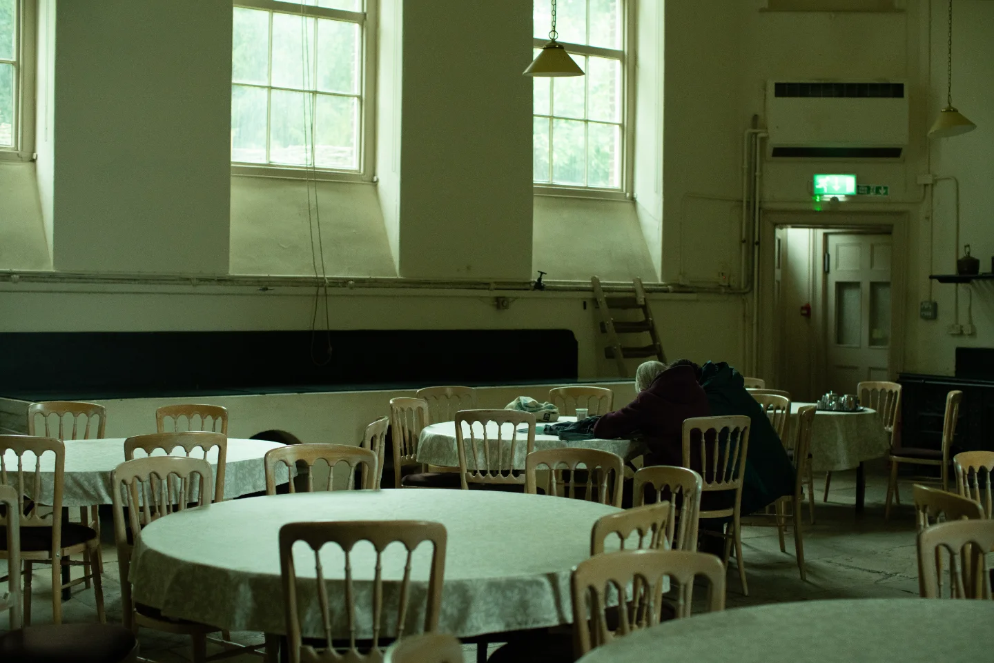 Round tables and white chairs wait beneath tall windows in a pale historic hall.