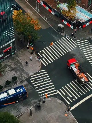 An overhead view of a New York intersection with striped crosswalks, pedestrians, a blue bus, and a red work truck beside an autumn tree.