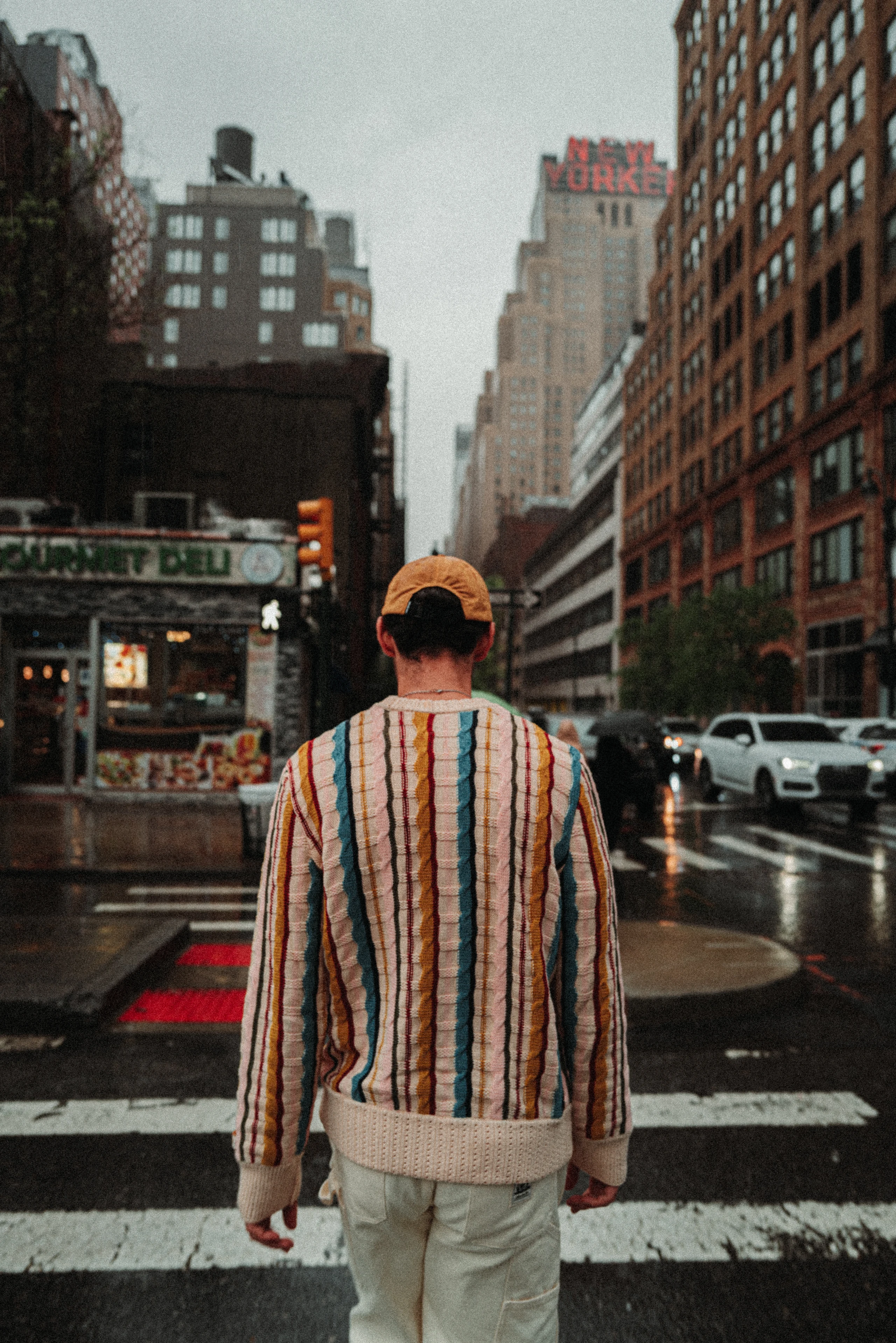 Person in a striped sweater and tan cap stands at a rainy Manhattan crosswalk with the New Yorker Hotel sign in the distance.