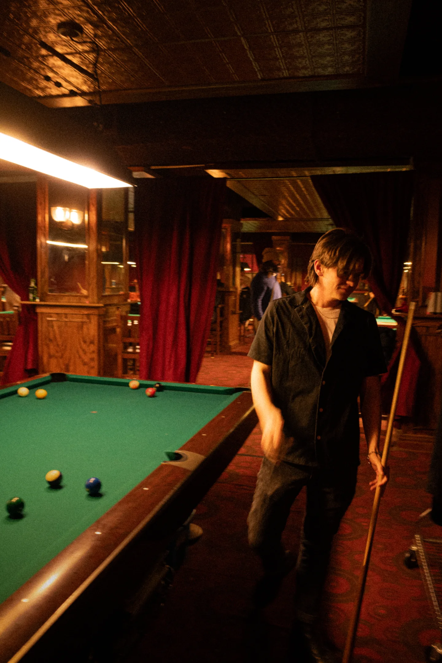 A player leans over the far side of a green pool table beneath amber light.