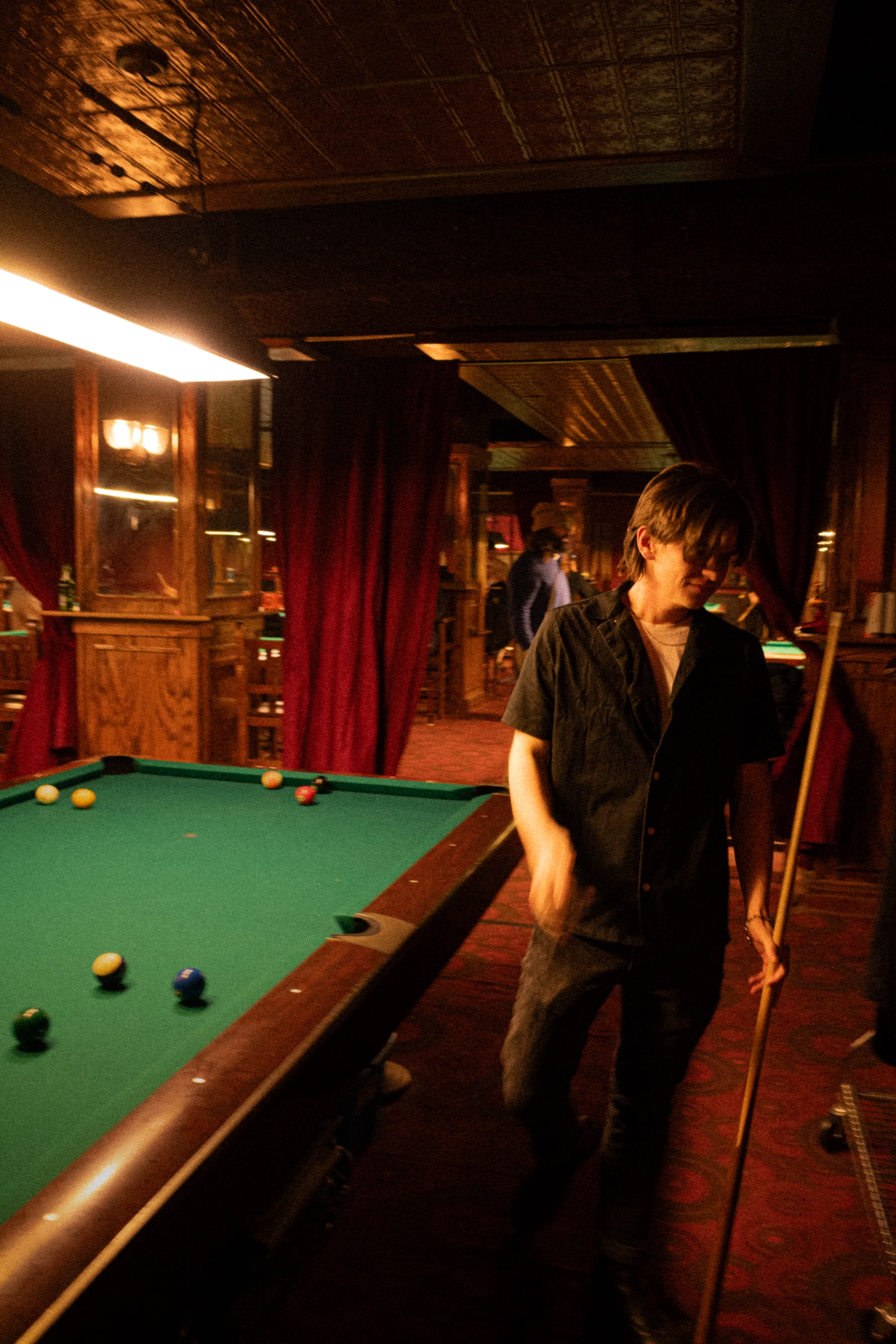 A player leans over the far side of a green pool table beneath amber light.