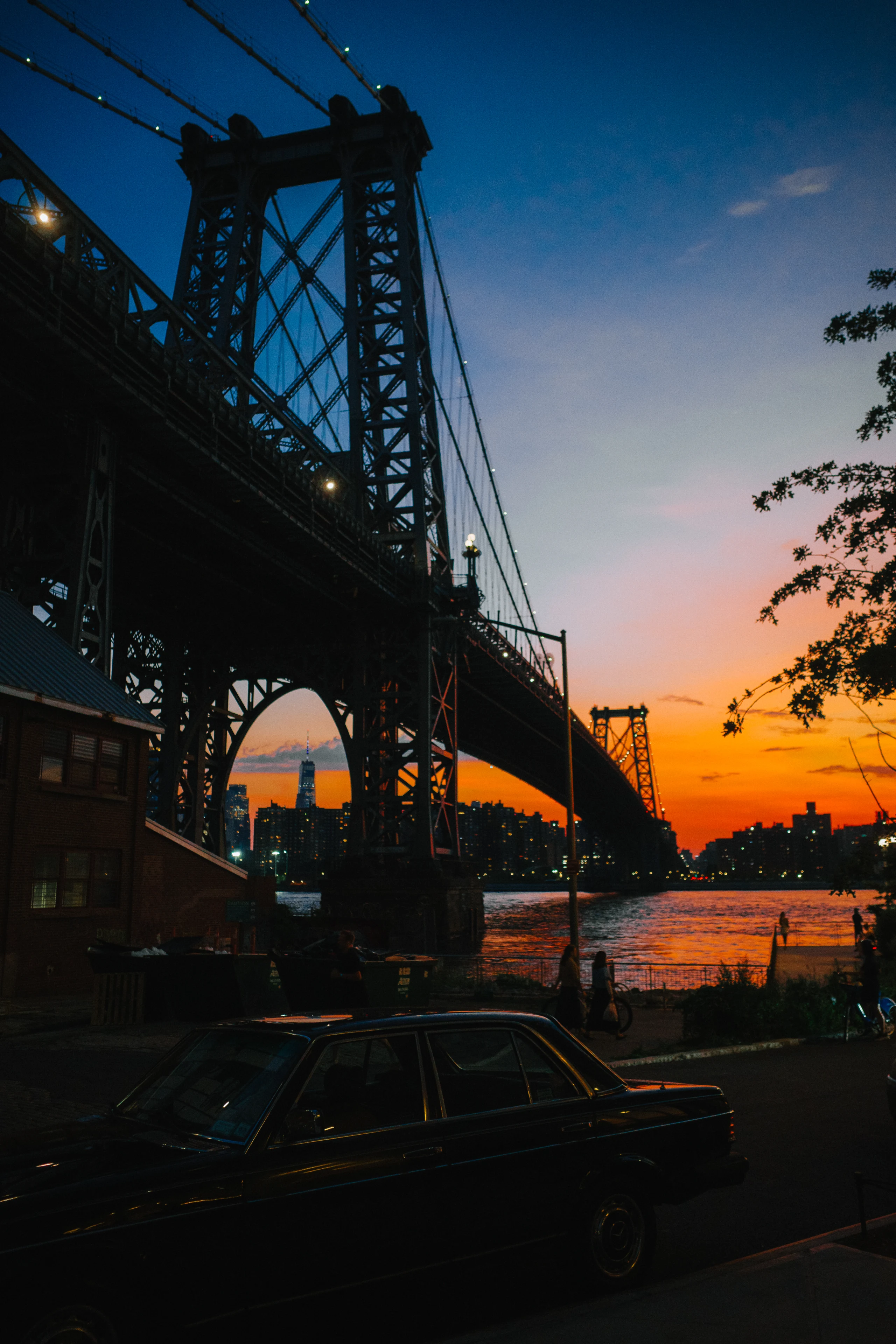 Williamsburg Bridge silhouetted against a blue and orange sunset above the East River, with a black car parked in the foreground.